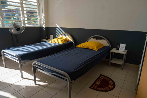Chambre simple et lumineuse avec deux lits une place équipés de draps bleus et d’oreillers jaunes. Chaque lit est accompagné d’une petite table de chevet sur laquelle sont posés une lampe et un livret. Un ventilateur mural est fixé près de la fenêtre à persiennes laissant entrer la lumière du jour. Le sol est carrelé et un petit tapis semi-circulaire est placé entre les deux lits. L’ensemble donne une impression d’ordre, de propreté et de simplicité.
