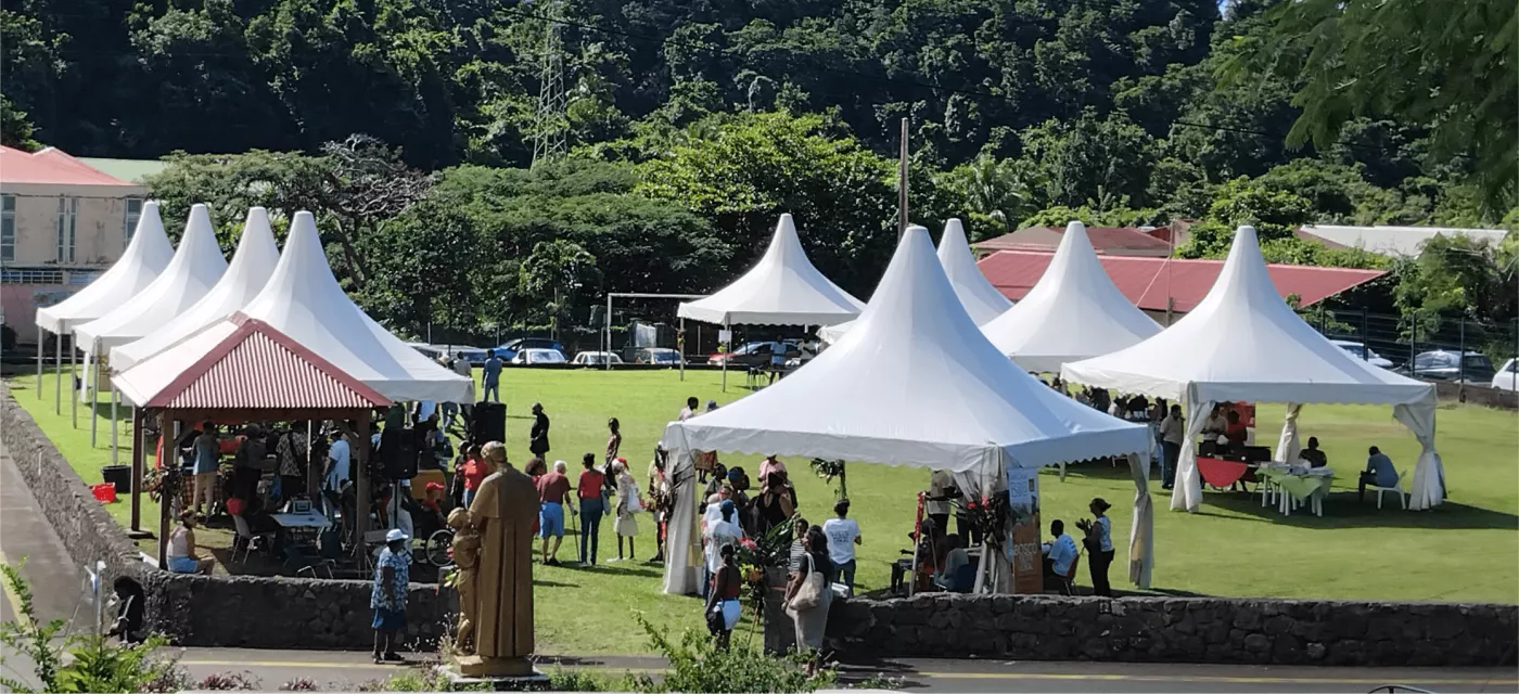 Vue d’un événement en plein air sur un grand espace vert entouré de montagnes tropicales. Plusieurs tentes blanches de type chapiteau sont installées, chacune abritant des groupes de personnes. Une statue en bronze de Don Bosco est visible au premier plan. L’ambiance est conviviale, avec des visiteurs et participants circulant entre les stands. Le ciel est bleu et dégagé. Quelques bâtiments aux toits rouges se trouvent à l’arrière-plan. 