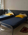 Chambre simple et lumineuse avec deux lits une place équipés de draps bleus et d’oreillers jaunes. Chaque lit est accompagné d’une petite table de chevet sur laquelle sont posés une lampe et un livret. Un ventilateur mural est fixé près de la fenêtre à persiennes laissant entrer la lumière du jour. Le sol est carrelé et un petit tapis semi-circulaire est placé entre les deux lits. L’ensemble donne une impression d’ordre, de propreté et de simplicité.